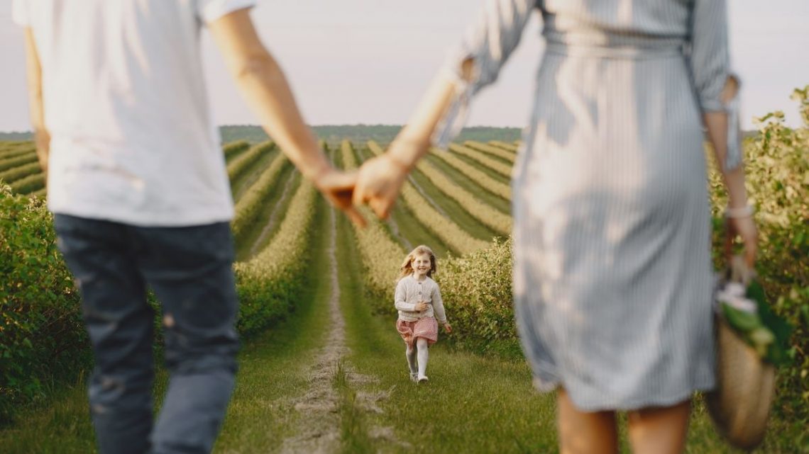 A family holds hands with their child in a beautiful, green field under a clear sky.